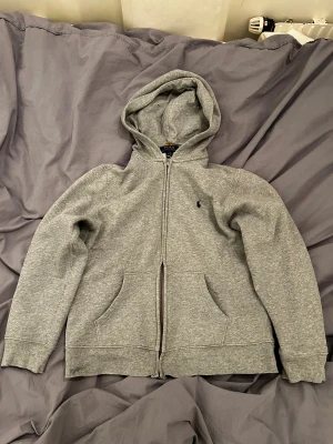 Grå hoodie från Polo Ralph Lauren - Snygg grå hoodie från Polo Ralph Lauren med dragkedja och huva. Tröjan har långa ärmar, två fickor fram och den klassiska lilla Polo-loggan broderad på bröstet. Mjuk insida i bomullsmix som gör den extra skön. Har en liten defekt i luvan (se bild 4) Strl L (14-16) OBS skriv till mig innan köp.