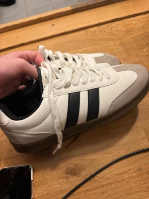 Adidas Samba vita sneakers - Fräscha Adidas Samba sneakers i vitt skinn med svarta klassiska ränder på sidorna och beige mockadetaljer vid tån. Skorna har rund tå, platt sula och snörning. Perfekt för dig som gillar retrostil och sportig vibe.