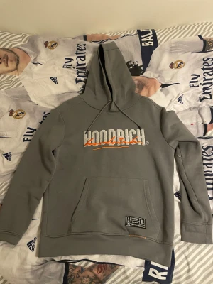 Grå hoodie från Hoodrich med tryck - Säljer en grå hoodie från Hoodrich i storlek M. Tröjan har stor huva, magficka och tryck både fram och bak med vit och orange text. Mjuk insida och klassisk passform, perfekt för chill dagar. Snygg detalj med etikett nertill.