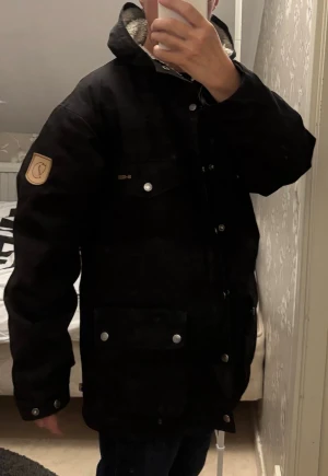 Svart parkajacka från Fjällräven - Svart parkajacka från Fjällräven med G-1000-tyg, mysigt teddyfoder i huvan och flera fickor med tryckknappar. Klassisk patch på ärmen och justerbara ärmslut. Perfekt för kalla dagar och snygg streetstil.
