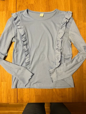 Ljusblå långärmad topp med volanger - Supersöt ljusblå långärmad topp från Indiska med volangdetaljer framtill. Materialet är mjukt och har en lätt struktur som ger en skön känsla. Perfekt för dig som gillar romantiska detaljer och vill ha något som sticker ut lite extra.