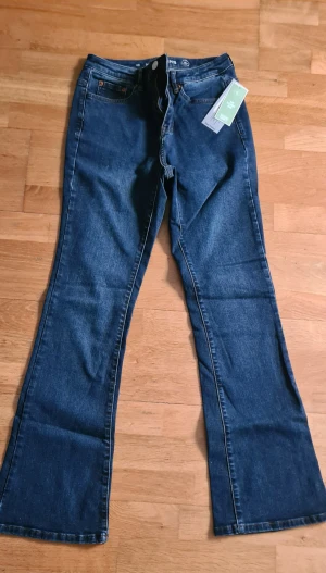 Blå bootcut jeans från KappAhl - Snygga mörkblå jeans från KappAhl i modellen Susan, med klassisk bootcut och hög midja. Jeansen har orangea sömmar, fem fickor och är tillverkade i mjukt bomullsdenim. Perfekta för dig som gillar retrovibbar och bekväm passform.