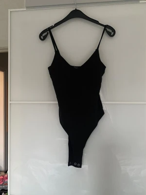 Svart bodysuit från Stradivarius XS - Svart bodysuit från Stradivarius i storlek XS. Modellen är ärmlös med smala axelband och v-ringad hals. Bodyn har tryckknappar i grenen för enkel på- och avtagning. Materialet är mjukt och stretchigt, perfekt för att skapa en clean och snygg siluett.