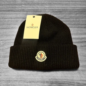 Moncler mössa - Svart ribbstickad mössa från Moncler med klassisk uppvikt kant och broderad Moncler-logga framtill. Snygg och enkel design som ger en clean look, perfekt för dig som gillar exklusiva märken. Materialet är mjukt och värmande ull.