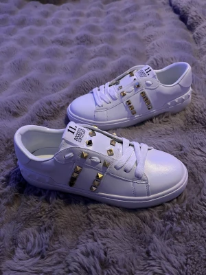 Valentino Rockstud Untitled vita sneakers - Snygga vita Valentino Garavani Rockstud Untitled sneakers med platt sula och rund tå. Skorna har coola guldfärgade nitar på ovansidan och sidorna, samt vita snören. Tillverkade i skinn för en lyxig känsla och stilren look. Perfekta för dig som gillar statement-detaljer.