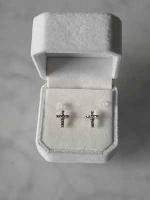 Silvriga korsörhängen - Ett par stilrena örhängen i form av kors, dekorerade med glittrande strass. De är silvriga och har en minimalistisk design som ändå sticker ut tack vare de små stenarna. Perfekta för dig som gillar detaljer med lite edge.