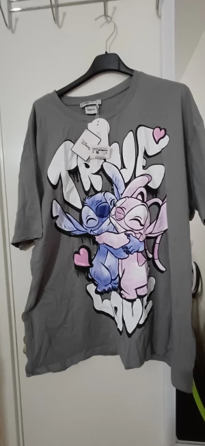 Disney Stitch t-shirt grå loose fit - Grå t-shirt från FB Sister med Disney Stitch och Angel-tryck framtill. Loose fit med korta ärmar och rund hals. Färgglada detaljer i blått, rosa och vitt samt hjärtan. Perfekt för dig som älskar söta och lekfulla motiv.