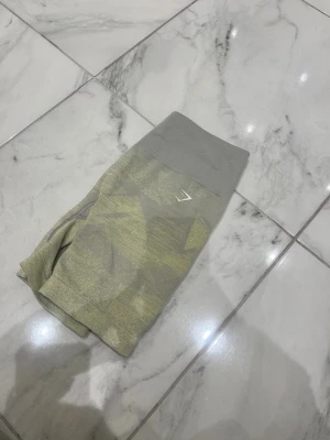 Grå-gula träningsshorts från Gymshark - Säljer ett par grå-gula seamless träningsshorts från Gymshark med hög midja och camo-inspirerat mönster. Stretchigt material som sitter tight och bekvämt, perfekt för gymmet eller andra sportaktiviteter. Diskret Gymshark-logga framtill.