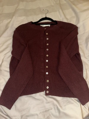 Vinröd kofta från Zara med knappar - Mysig vinröd kofta från Zara i storlek S. Koftan är stickad med rund hals och har pärlemorskimrande knappar framtill. Långärmad och med ribbade muddar vid ärmslut och nederkant. Perfekt för lager-på-lager under hösten och vintern.
