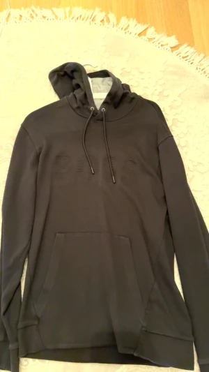 Svart hoodie från BOSS med broderad logga - Svart hoodie från BOSS med broderad logga framtill. Klassisk känguruficka, dragsko i huvan och mjukt material. Perfekt för dig som gillar stilren streetwear med premiumkänsla.