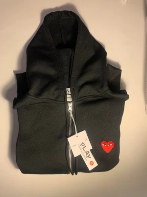 Svart hoodie Comme des Garçons Play - Svart hoodie från Comme des Garçons Play med dragkedja och huva. Tröjan har ett ikoniskt rött hjärta med ögon broderat på bröstet. Tillverkad i mjuk bomull och har ribbade muddar vid ärmslut och nederkant. Perfekt för en chill och stilren look.
