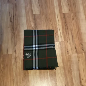 Rutig halsduk från Burberry London - Snygg halsduk från Burberry London med klassiskt rutigt mönster i mörkgrön, svart, vit och röd. Broderad logga i ena hörnet och fransar i kanterna. Perfekt accessoar för att lyfta din stil med en lyxig touch.