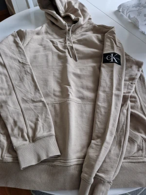 Beige hoodie från Calvin Klein  XL - Säljer en beige hoodie från Calvin Klein Jeans i storlek XL. Tröjan har huva med snörning, ribbade muddar och en stor CK-logga broderad på ärmen. Materialet är bomull och passformen är normal. 