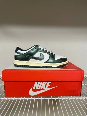 Nike Dunk Low vit och mörkgrön - Nike Dunk Low sneakers i vit och mörkgrön färg med klassisk snörning och rund tå. Ovandelen är i skinn med perforerad tåbox och gröna detaljer runt hela skon. Sulan är platt och har en retro off-white ton. Levereras med extra gröna skosnören och originalkartong.