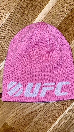 Äkta UFC merch pink beanie / rosa mössa - Rosa mössa med vit UFC logga framtill. En liten UFC etikett finns på sidan. Knappt använt.. Mössan är stickad och tillverkad i 100% akryl, vilket gör den mjuk och skön att bära. Perfekt för dig som gillar sportiga accessoarer och vill sticka ut med färg.