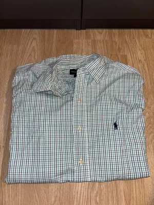 Rutig skjorta från Ralph Lauren M - Perfekt att ge din bror, pappa kille eller dig själv som en julklapp. Snygg rutig skjorta från Ralph Lauren, modell Blaire, i storlek M men passar även L. Skjortan är vit med gröna och mörkblå rutor, har klassisk krage och knappar framtill. Tillverkad i 100% bomull och har lång ärm samt broderad logga på bröstet.