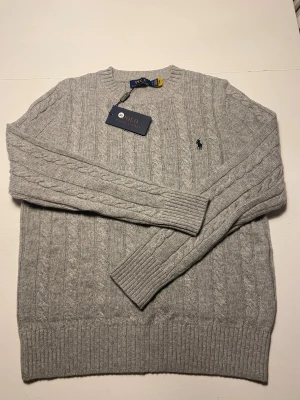 Grå stickad tröja från Polo Ralph Lauren - Snygg grå kabelstickad tröja från Polo Ralph Lauren med rund hals och diskret broderad logga på bröstet. Tröjan har långärmade ribbade muddar och är tillverkad i mjuk ull, perfekt för kyliga dagar.