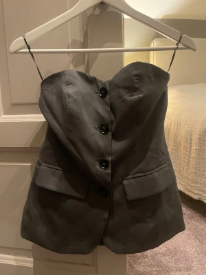 Grå tubtopp med knappar från Pieces - Säljer min ljusgrå tailored tubtopp från Pieces! Toppen har en snygg bandeauform med stora svarta knappar framtill, är i strl XS och döljer pga att den inte kommer till användning❣️