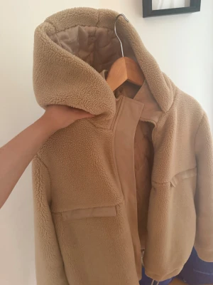 Beige teddyjacka med huva från Saint - Mysig teddyjacka i beige från Saint, storlek S. Jackan har en stor huva, två fickor framtill och stängs med dragkedja. Utsidan är fluffig och insidan är quiltad för extra värme. Perfekt för kyliga dagar och har en clean, modern look.