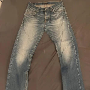 Blå jeans från Nudie Jeans Co - Säljer ett par klassiska blå jeans från Nudie Jeans Co med snygga slitningar och orangea sömmar. Jeansen har fem fickor och raka ben, perfekt för en avslappnad stil. Midjan är normal och passformen är straight. Finns en defekt som är med på sista bilden. Nyansen kan skilja sig lite åt. 