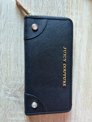 Svart plånbok från Juicy Couture - Snygg svart plånbok från Juicy Couture med guldfärgade detaljer och logotyp på framsidan. Plånboken har flera fack, dragkedja runtom och insida med blått tyg och kronmönster. Materialet är strukturerat syntet med en stilren och modern look.