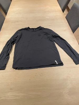 Grå långärmad tröja med logga - Grå tunn långärmad tröja från Sailracing i storlek small. Tröjan har en rund hals och ett broderat logomärke på bröstet. Tillverkad i mjuk bomull, perfekt för chill dagar. Enkel och stilren design utan mönster eller extra detaljer. Inga skador på tröjan men haft den ganska länge