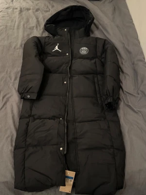 PSG lång dunjacka - Säljer en svart lång dunjacka från Jordan x PSG med huva och vita loggor på bröstet. Jackan har dragkedja och tryckknappar framtill, samt elastiska muddar vid ärmslut. Perfekt för kalla dagar och har en riktigt clean look.