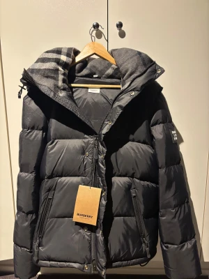 Svart pufferjacka från Burberry - Svart pufferjacka från Burberry med ikonisk rutig huva i grå toner. Jackan har dragkedja och knappar framtill, två sidofickor med dragkedja och en loose passform. Materialet är polyester och dun, perfekt för kalla dagar. Stilsäker och klassisk design med Burberry-logga på ärmen. Man kan även göra om den till en väst. STORLEK M passar även liten L 