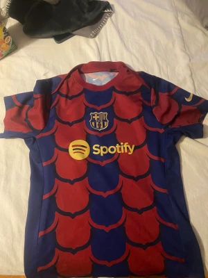Barcelona Nike Dri-FIT fotbollströja S - Barcelona fotbollströja från Nike med Dri-FIT material. Tröjan har korta ärmar och ett unikt mönster i mörkblått och rött, med klubbmärket och Spotify-logga på bröstet samt Nike-logga i gult på ärmen. Perfekt för dig som älskar Barca och vill sticka ut på planen.