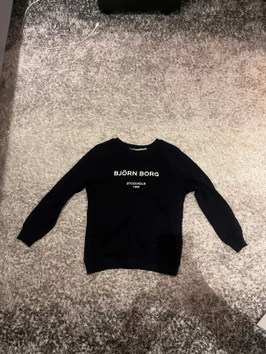 Marinblå Björn Borg sweatshirt - Svart sweatshirt från Björn Borg med vit text på bröstet. Tröjan har rund hals, långa ärmar och ribbade muddar vid ärmslut och nederkant. Perfekt för chill dagar och enkel att matcha med jeans eller joggers.