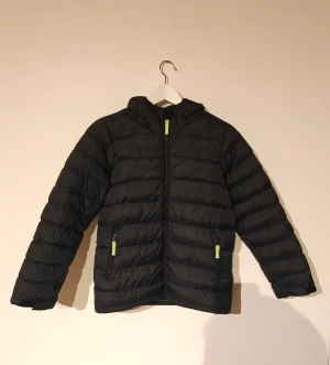 Tuxer vinterjacka  - Svart pufferjacka från Tuxer med huva och gröna detaljer på dragkedjorna. Jackan har quiltad design, två sidofickor med dragkedja och är tillverkad i polyamid och polyester. Perfekt för kyliga dagar och har en snygg, sportig look. Hängaren är sönder och har ett hål vid ryggen som är uppsytt och ett där luvan knäpps fast.