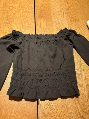 Svart offshoulder topp från BikBok - Svart offshoulder topp från BikBok i storlek S. Toppen har långa ärmar och är smockad både vid axlar, midja och ärmslut för en snygg och figurnära passform. Materialet är mjukt och lätt, perfekt för att skapa en cool och trendig look.