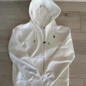 Vit hoodie från Polo Ralph Lauren - Vit hoodie från Polo Ralph Lauren med broderad marinblå logga på bröstet. Tröjan har huva med snörning, dragkedja framtill och två fickor. Tillverkad i mjuk bomull, perfekt för chill dagar. Klassisk och clean look som alltid funkar.