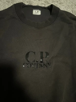Svart sweatshirt från C.P. Company - Svart sweatshirt från C.P. Company med stilrent tryck på bröstet. Tröjan har rund halsringning och långa ärmar. Materialet är mjuk bomull och passformen är normal. Perfekt för dig som gillar clean streetwear och vill ha något enkelt men snyggt. St M men passar även S