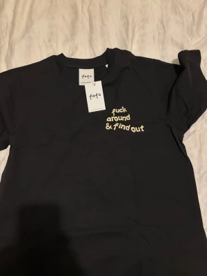 Fafo  - Svart t-shirt med trycket ”fuck around & find out” framtill och en smiley med stjärnor till ögon baktill. T-shirten har även en etikett.