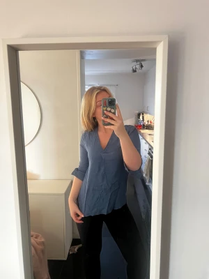 Blå v-ringad blus med trekvartsärm - Säljer en stilren blå blus med v-ringning och trekvartsärmar. Blusen har en mjuk och skön passform, perfekt att matcha med jeans eller kjol. Snygg detalj med knapp vid ärmslutet. Passar dig som gillar enkel men trendig stil.