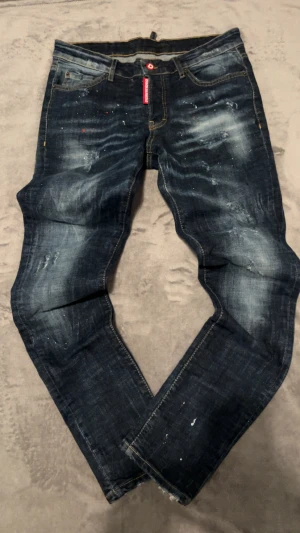 Dsquared2 Slim Jeans blå - Snygga Dsquared2 Slim Jean i mörkblå tvätt med slitningar och färgstänk för en cool, edgy look. Klassisk femficksmodell med kontrastsömmar och slim passform. Tillverkade i Italien av högkvalitativt jeanstyg. Perfekt för dig som gillar att sticka ut lite och pris kan diskuteras vid snabb affär!