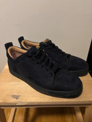 Mörkblå Louboutin sneakers mocka - Snygga mörkblå sneakers från Louboutin i mjuk mocka med klassisk röd sula och broderad logga på hälen. Skorna har mörkblåa snören, rund tå och beige innersida i skinn. Perfekta för dig som vill ha en clean men lyxig look. Kan gå ned i pris vid snabb affär