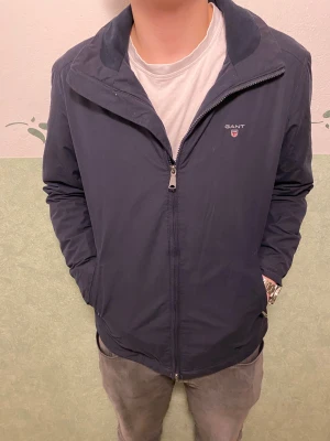 Mörkblå jacka från GANT - Snygg mörkblå vindjacka från GANT med klassisk ståkrage och diskret logga på bröstet. Jackan har dragkedja framtill, två sidofickor och är tillverkad i ett lätt och slitstarkt syntetmaterial. Perfekt för dig som gillar stilren och enkel design.