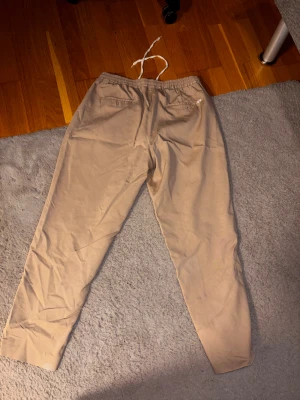 Beige chinos från Selected Homme W36 L32 - Snygga beige chinos från Selected Homme med elastisk midja och vit snörning. Klassisk rak passform och tillverkade i mjukt bomullsmaterial. Perfekta för en chill och stilren look, passar dig som gillar enkel och clean stil.