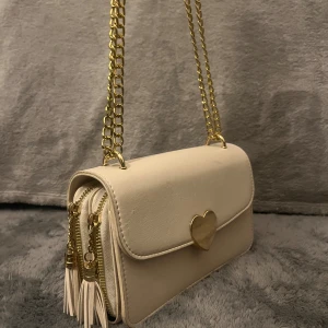 Beige axelväska med hjärt-detalj - Supersöt beige axelväska i skinnimitation med guldfärgad kedja och hjärtformat spänne framtill. Väskan har två dragkedjefack med tofsar och stilren, rektangulär form. Perfekt skick