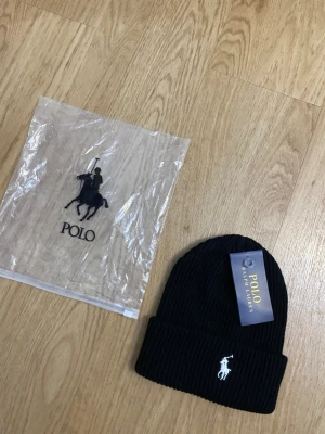 Svart ribbad mössa från Polo Ralph Lauren - Svart ribbstickad mössa från Polo Ralph Lauren med klassisk broderad logga i vitt framtill. Mössan har uppvikt kant och är tillverkad i mjukt material som håller dig varm. Perfekt accessoar för att lyfta din stil under kyliga dagar.
