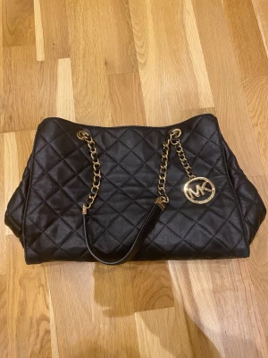 Svart quiltad axelväska Michael Kors - Väska från Michael Kors i quiltat skinn med guldfärgade kedjehandtag och MK-logga. Väskan har flera fack, dragkedja och detaljer i guld. Lite defekter/slitage på undersidan (visas på bild), annars bra skick och fungerar väl. 