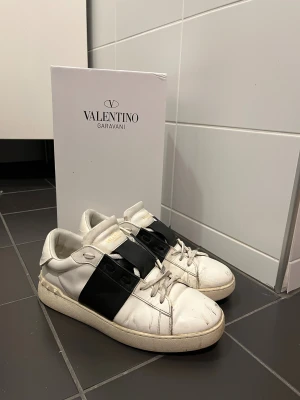 Valentino Garavani Open sneakers vit/svart - Säljer ett par Valentino Garavani Open sneakers i vitt skinn med bred svart läderdetalj över sidan. Skorna har snörning, rund tå och platt sula med ikoniska nitar baktill. Perfekt för dig som gillar lyxig stil. Skorna är i bra skick trots användning och har då några defekter därav pris, det är slitningar fram på tån, någon slitning under skon men inga hål och går jättebra att använda. Sulan är lite gul men går att fixa genom att använda vit sul penna vilket går att köpa!