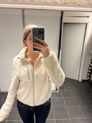 Vit teddyjacka med dragkedja - Säljer en fluffig vit teddyjacka med hög krage och hel dragkedja framtill. Jackan har en bröstficka med dragkedja i blankt tyg som kontrast. Perfekt för dig som gillar mysiga och trendiga jackor med chill vibe.