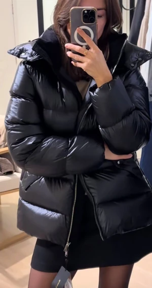 Svart pufferjacka med huva - Säljer en svart, glansig pufferjacka med stor huva och dragkedja framtill. Jackan har en loose passform och är quiltad med breda kanaler. Perfekt för kalla dagar och har en clean, modern look som funkar till många stilar.