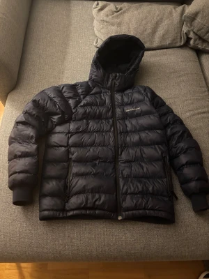 Mörkblå Peak Performance pufferjacka - Säljer en mörkblå pufferjacka från Peak Performance med huva och dragkedja framtill. Jackan har quiltad design, två sidofickor med dragkedja och ribbade muddar vid ärmslut. Perfekt för kalla dagar och har en clean, sportig look.