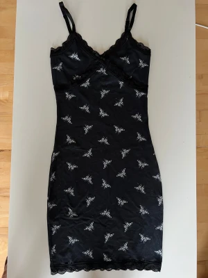 Svart kort klänning med fjärilar - Supersöt svart kort klänning med vita fjärilar i tribalstil. Klänningen har smala axelband, spetsdetaljer vid urringningen och nederkanten samt en liten rosett framtill. Figurnära passform och mjukt material som känns skönt mot huden.