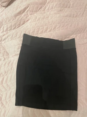 Svart tight kortkjol - Säljer en svart, stilren och tight kortkjol med bred linning. Kjolen har en enkel och klassisk design utan mönster eller detaljer, perfekt för att matcha med olika toppar. Materialet känns mjukt och stretchigt, vilket gör den bekväm att bära hela dagen.