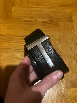 Svart skärp Tom Ford T Icon - Säljer ett Tom Ford T Icon bälte i svart skinn med en stilren T-formad metallspänne i silver. Bältet har exklusiv känsla och är perfekt för dig som gillar statement-accessoarer. Märket är diskret stämplat på insidan av bältet.